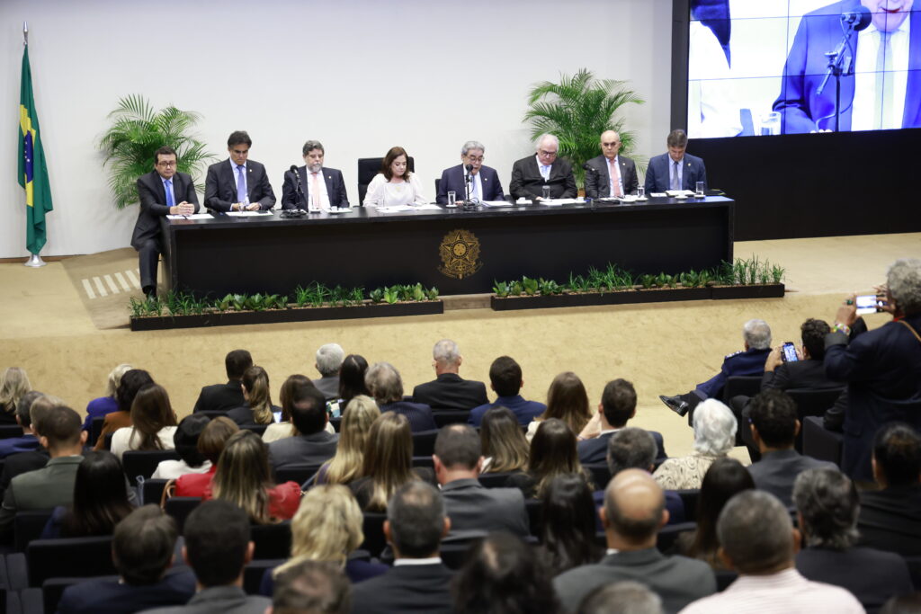 Imagem da solenidade em homenagem aos 20 anos do CSJT, realizada no plenário da Câmara dos Deputados. A mesa de honra é composta por oito autoridades, posicionadas atrás de uma bancada com o brasão da República. O auditório está cheio, com parte do público registrando o evento com celulares. Ao fundo, há bandeira do Brasil, plantas ornamentais e uma tela que transmite a cerimônia ao vivo.