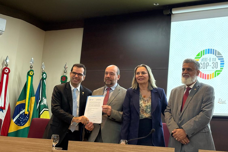 Ministros do TST posam para foto durante cerimônia de assinatura de documento, em evento institucional com temática relacionada à COP-30 e aos Objetivos de Desenvolvimento Sustentável (ODS). Ao fundo, há bandeiras e projeção do logotipo “ART-8 na COP-30”.