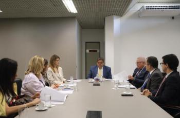 A imagem mostra um grupo de pessoas em reunião ao redor de uma mesa, com documentos à frente, em um ambiente corporativo.