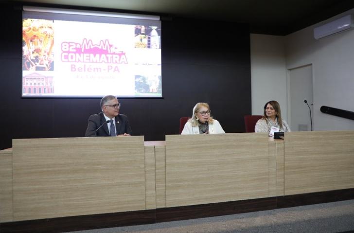 Fotografia em ambiente fechado que reúne em uma mesa oficial de evento duas mulheres e um homem.