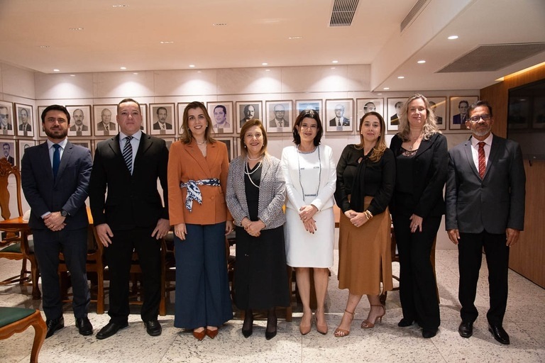 A imagem mostra um grupo de oito pessoas posando para a foto em um ambiente formal, possivelmente institucional.