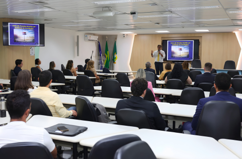 Técnicas para negociação marcam evento preparatório da Semana Nacional de Conciliação - CSJT2 6 Fotografia de uma palestra com algumas pessoas sentadas assistindo e um palestrante em pé ao lado de um banner.