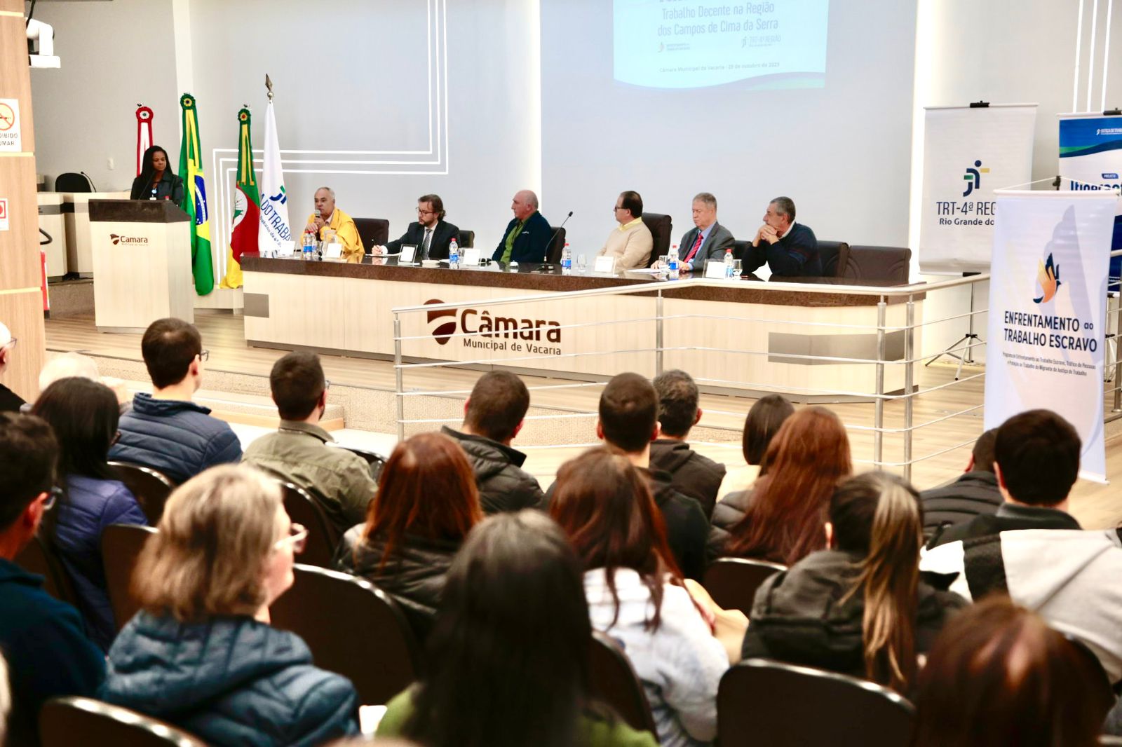 Todas as Notícias 1 Fotografia de uma mesa diretora e de um público sentado; ao lado alguns cartazes e acima deles um slide projetado.