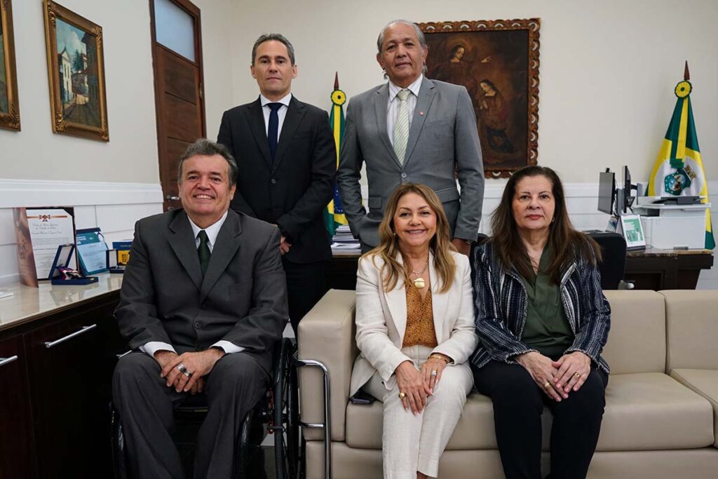 Fotografia com a presença de 5 pessoas, sendo 3 homens e 2 mulheres; as mulheres estão sentadas, um dos homens está em uma cadeira de rodas e os outros 2 estão em pé.