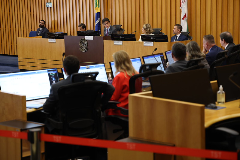 Foto do plenário da sessão do CSJT desta sexta-feira em um ângulo em diagonal, com o primeiro plano desfocado e alguns conselheiros e o presidente, ao centro e em segundo plano, com foco