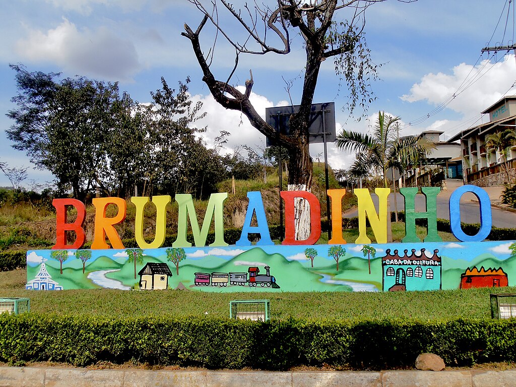 Portal da cidade de Brumadinho, com nome da cidade em letras coloridas sobre um muro com pinturas de igreja, casa, trem e casa da cultura