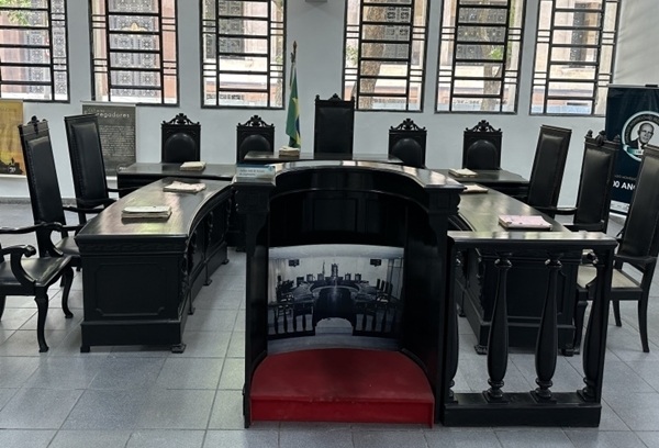 Fotografia de uma mesa com diversas cadeiras ao redor e janelas ao fundo.