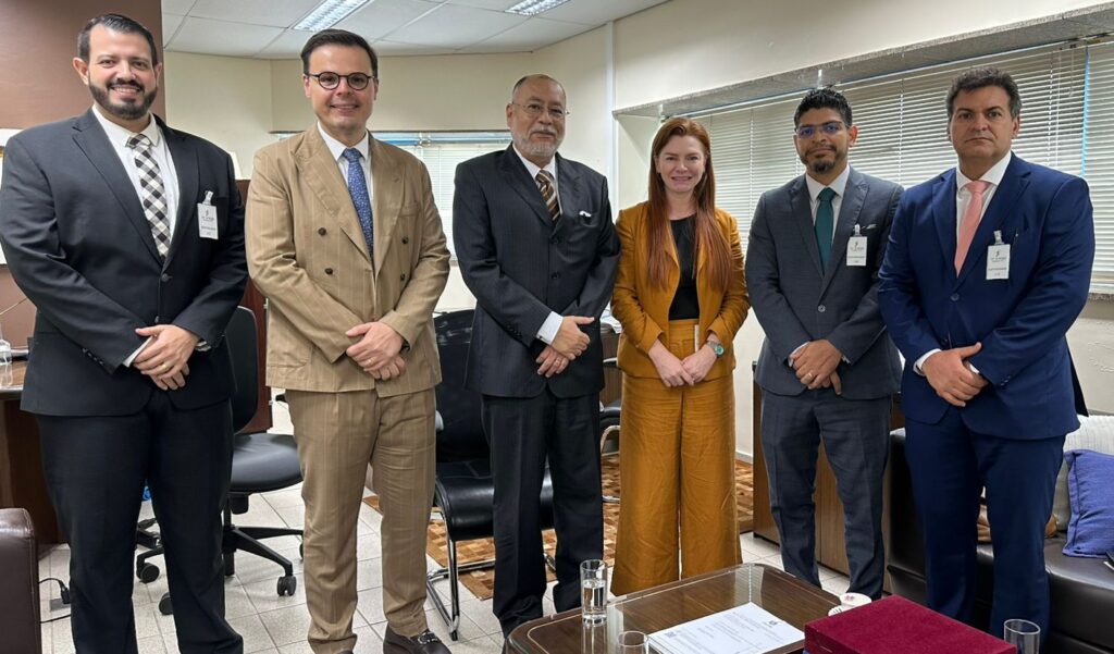 Fotografia com 6 pessoas em pé, em frente a uma mesa, sendo 5 homens e 1 mulher.