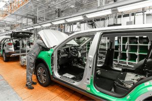 Todas as Notícias 2 A imagem mostra um operário uniformizado trabalhando na parte frontal de um veículo prata em uma linha de montagem automotiva moderna e iluminada.