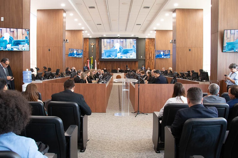 Todas as Notícias 3 Fotografia de uma sala de reunião, com bancadas diretivas e pessoas ao fundo assistindo.